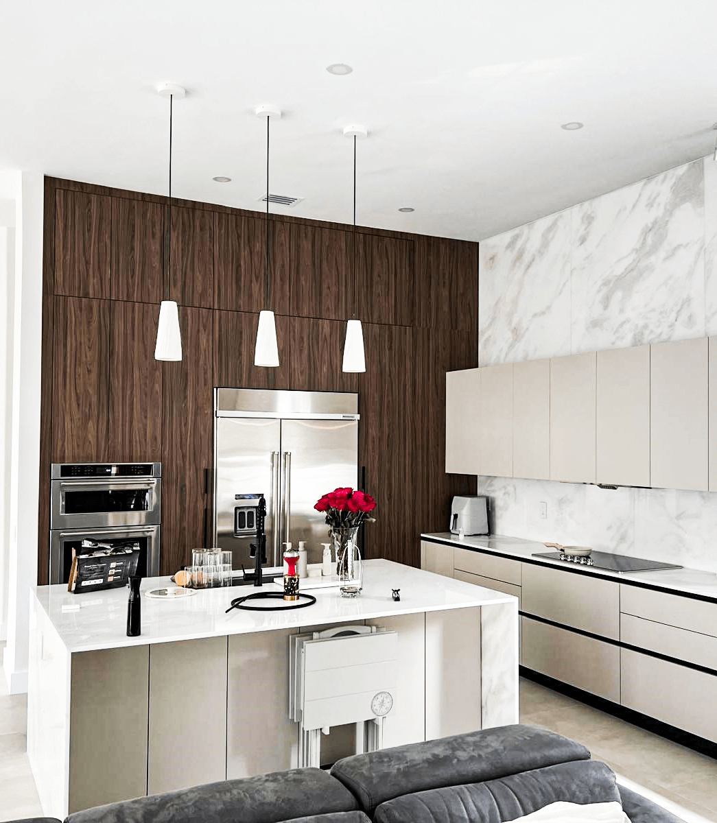 Modern kitchen featuring a marble countertop island, beige cabinetry, stainless steel appliances, and a wood-accent wall with pendant lighting. A vase with red flowers adds a touch of elegance to the space.