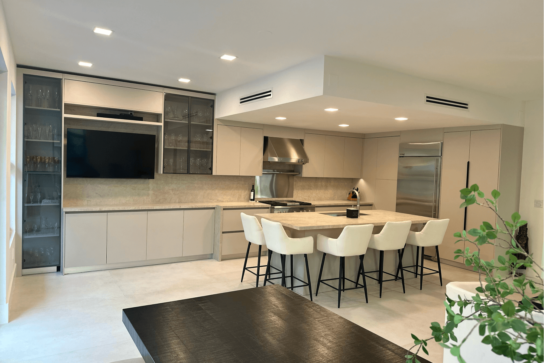 Modern kitchen with beige cabinets, built-in appliances, a large island with bar stools, and glass shelving for glassware