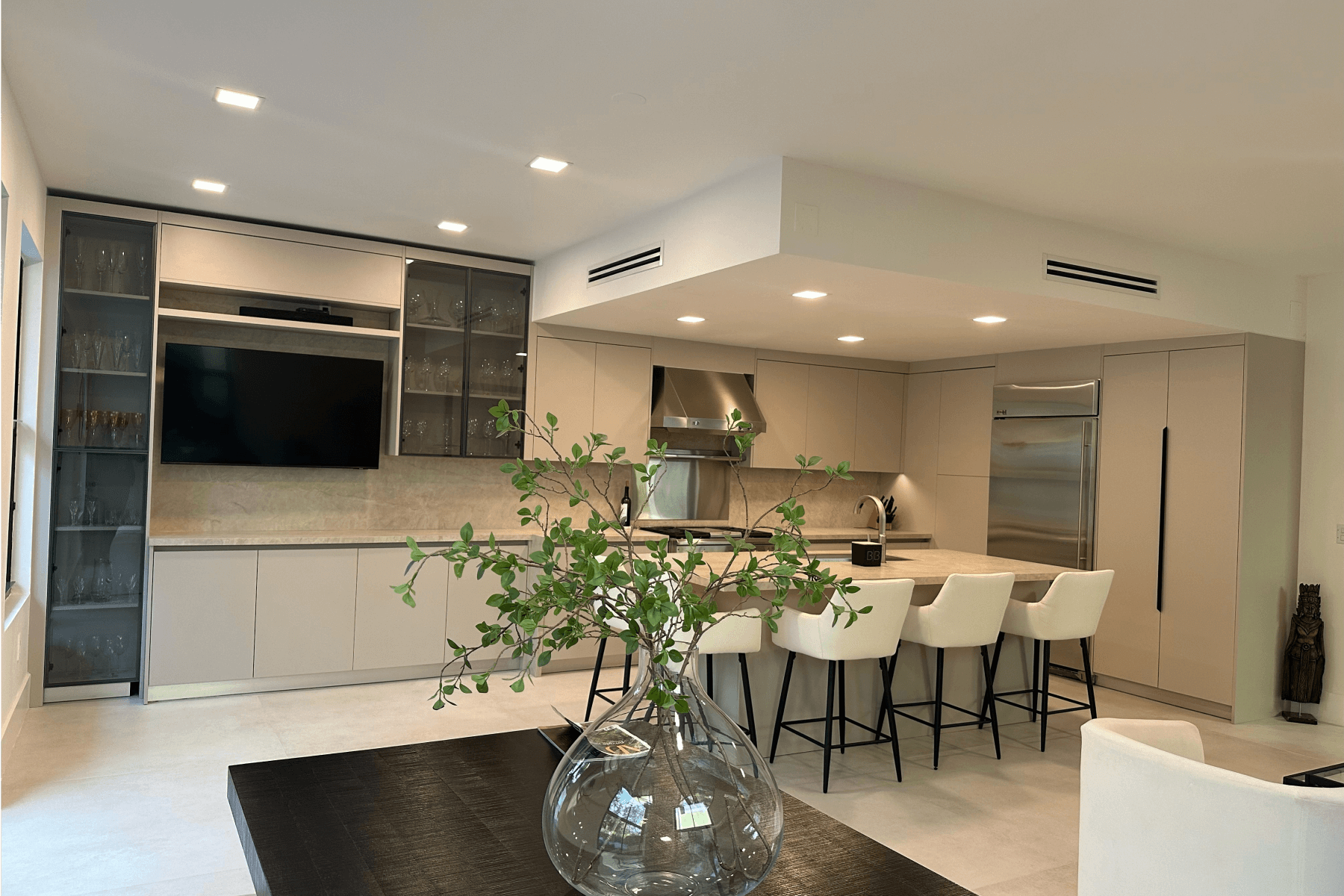 Kitchen with a modern design, featuring beige cabinetry, a center island with white bar stools, glass cabinets, and a decorative plant in a glass vase.