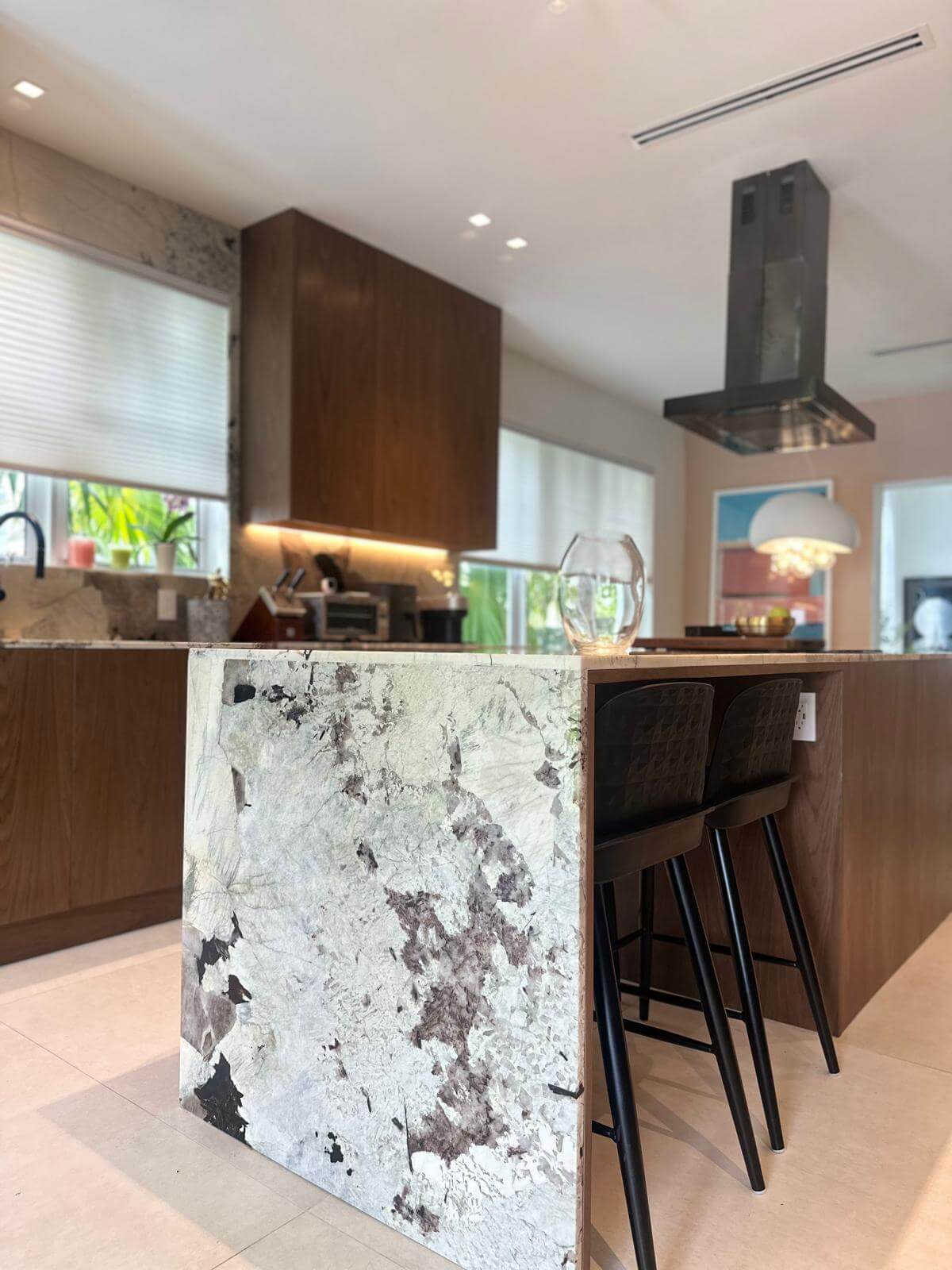 Modern kitchen with a sleek island featuring a striking marble waterfall countertop and wooden accents. Stylish black barstools complement the clean, contemporary design, while natural light highlights the rich wood cabinetry and modern range hood.