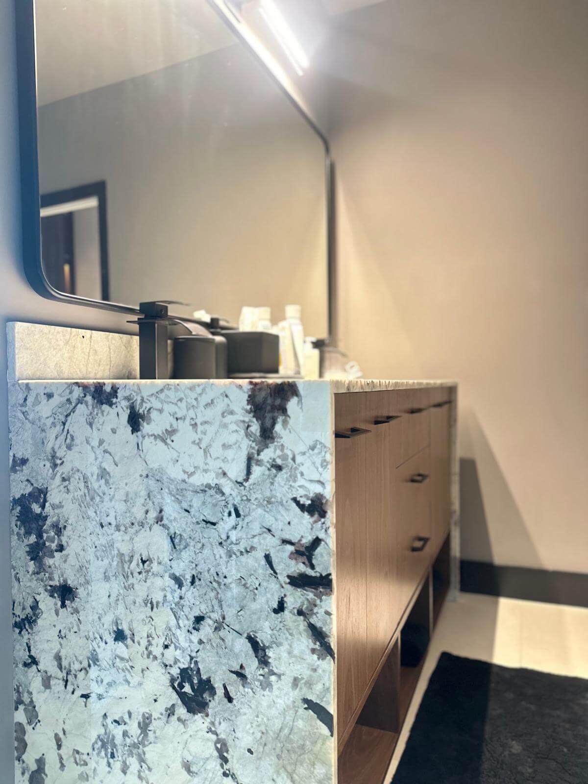 Modern bathroom featuring a luxurious marble countertop with bold veining and a wooden vanity with sleek, minimalist drawers. The large, illuminated mirror enhances the space, adding depth and elegance to the contemporary design.