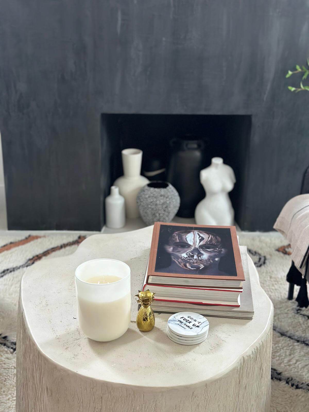 Stylish modern living room setup with a textured off-white coffee table featuring a candle, decorative objects, and a stack of artistic books. A black accent wall with a built-in niche holds a collection of minimalist vases and sculptures, adding depth and sophistication to the space.