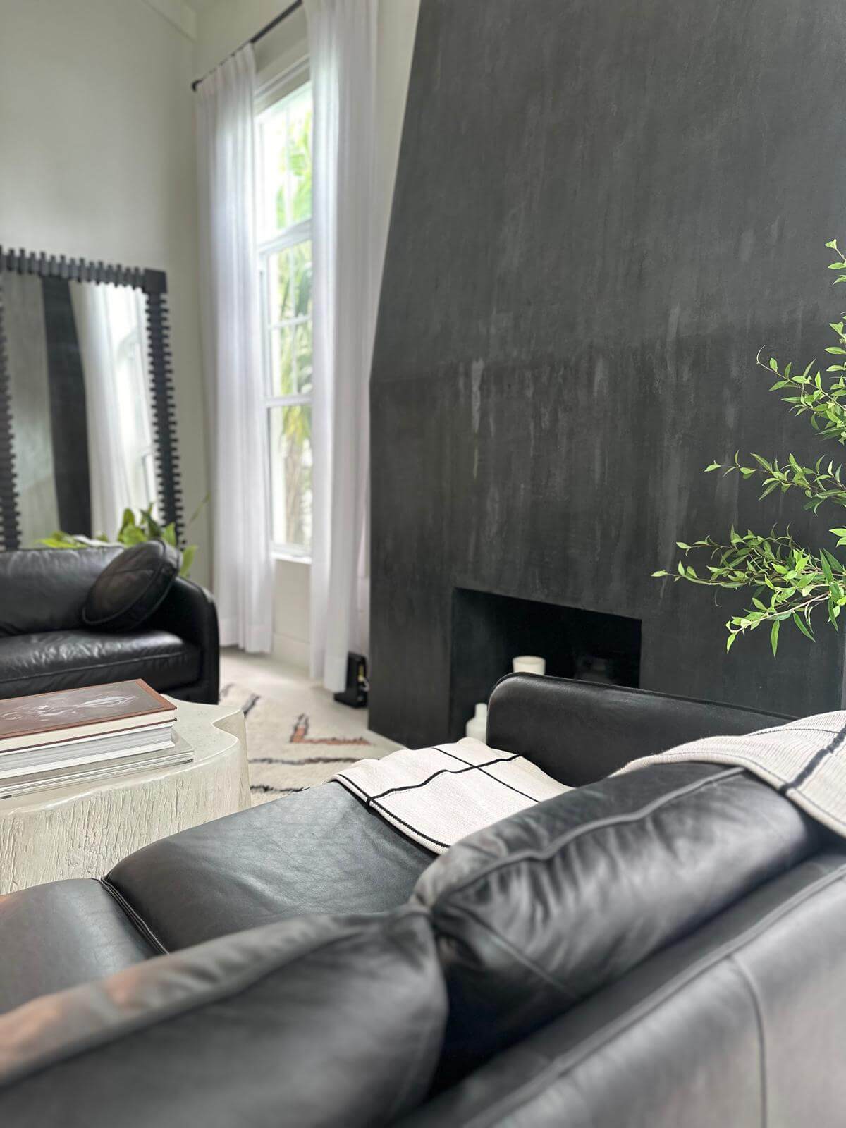 Modern living room featuring a bold black textured accent wall with a built-in niche, paired with luxurious black leather sofas. The space is softened by light, flowing curtains framing tall windows that let in natural light. A stylish white coffee table holds stacked books, adding a refined touch to this contemporary, cozy setting.