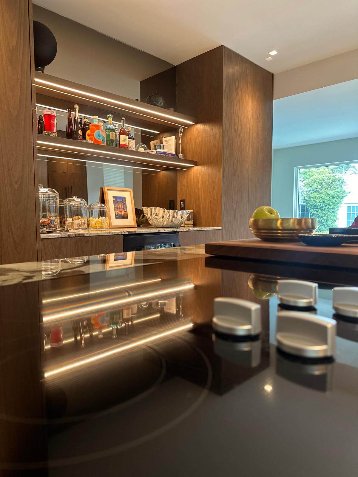 Modern kitchen space featuring a sleek black glass cooktop with reflective surfaces and metallic knobs. In the background, elegant wooden cabinetry with warm LED lighting highlights open shelves stocked with glassware, bottles, and decorative pieces. A polished brass bowl adds a stylish touch on the countertop, while a large window brings in natural light, enhancing the inviting and sophisticated atmosphere.